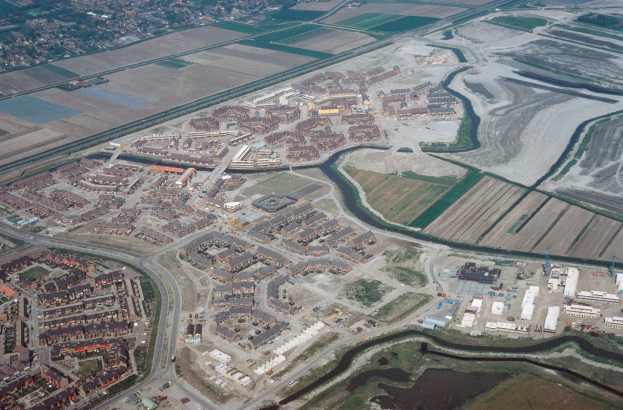 Luftaufnahme einer Stadt mit dichtem Gebäudeensemble, Straßen, Feldern, Bäumen und einem zentralen Reservoir, umgeben von städtischen und grünen Bereichen.