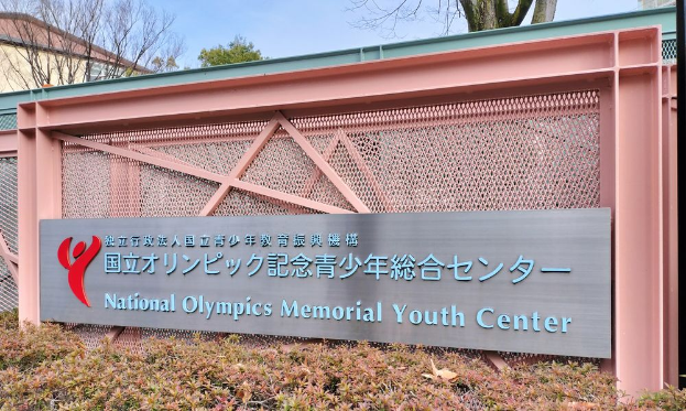 Das Gebäude des Nationalen Olympischen Memorial Youth Center mit einer Wandtafel, Pflanzen am Boden, Bäumen und Gebäuden im Hintergrund und einem klaren blauen Himmel darüber.