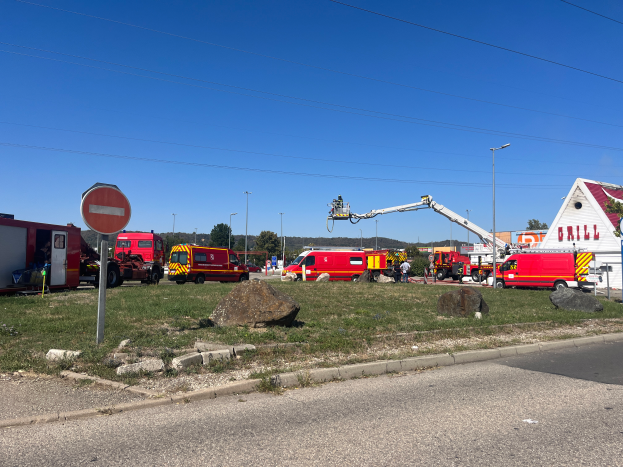Eine Gruppe von Feuerwehrautos steht auf der Seite einer Straße, umgeben von Gras, Steinen, einem Schild, einem Haus, Pfählen, einem Kran, Drähten, Bäumen und einem bewölkten Himmel, wahrscheinlich nach einem Brand an einer Tankstelle.
