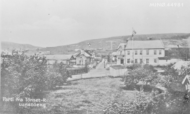Ein Schwarz-Weiß-Foto einer kleinen Stadt in Lunstöeng, Norwegen, circa 1900, das Gebäude, Bäume, Pfosten, Flaggen, einen Zaun, Berge und einen klaren Himmel zeigt.