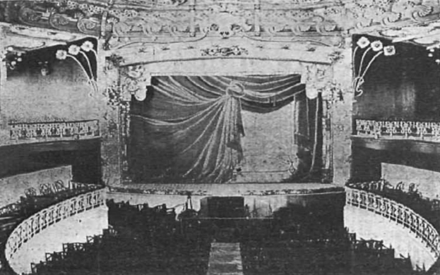 Schwarz-weiß-Foto des Auditoriums des Theatre Royal in London, das eine Bühne mit Vorhang, Geländer und ornamentale Wandgestaltung zeigt, mit ein paar sitzenden Menschen und Text unten.
