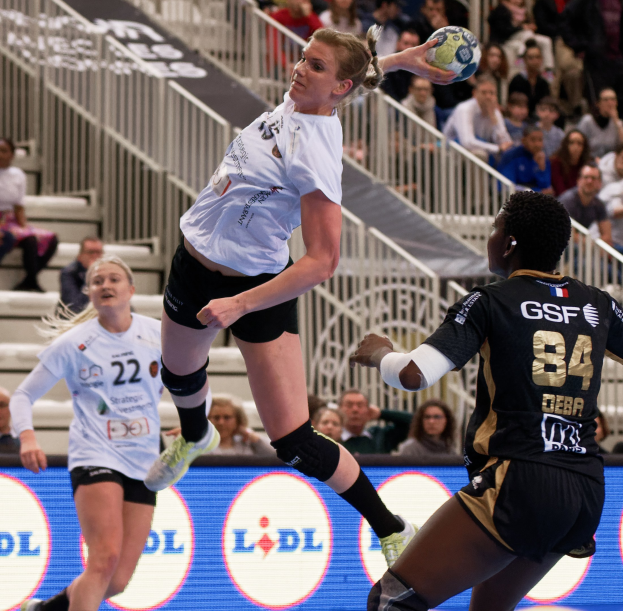 Eine Gruppe von Frauen, die Handball auf einem Platz spielt, mit einem Ball in der Luft, Zuschauern auf Treppen und Geländern im Hintergrund und einem "Handball-Weltmeisterschaft 2019 - Tag 2"-Schild.