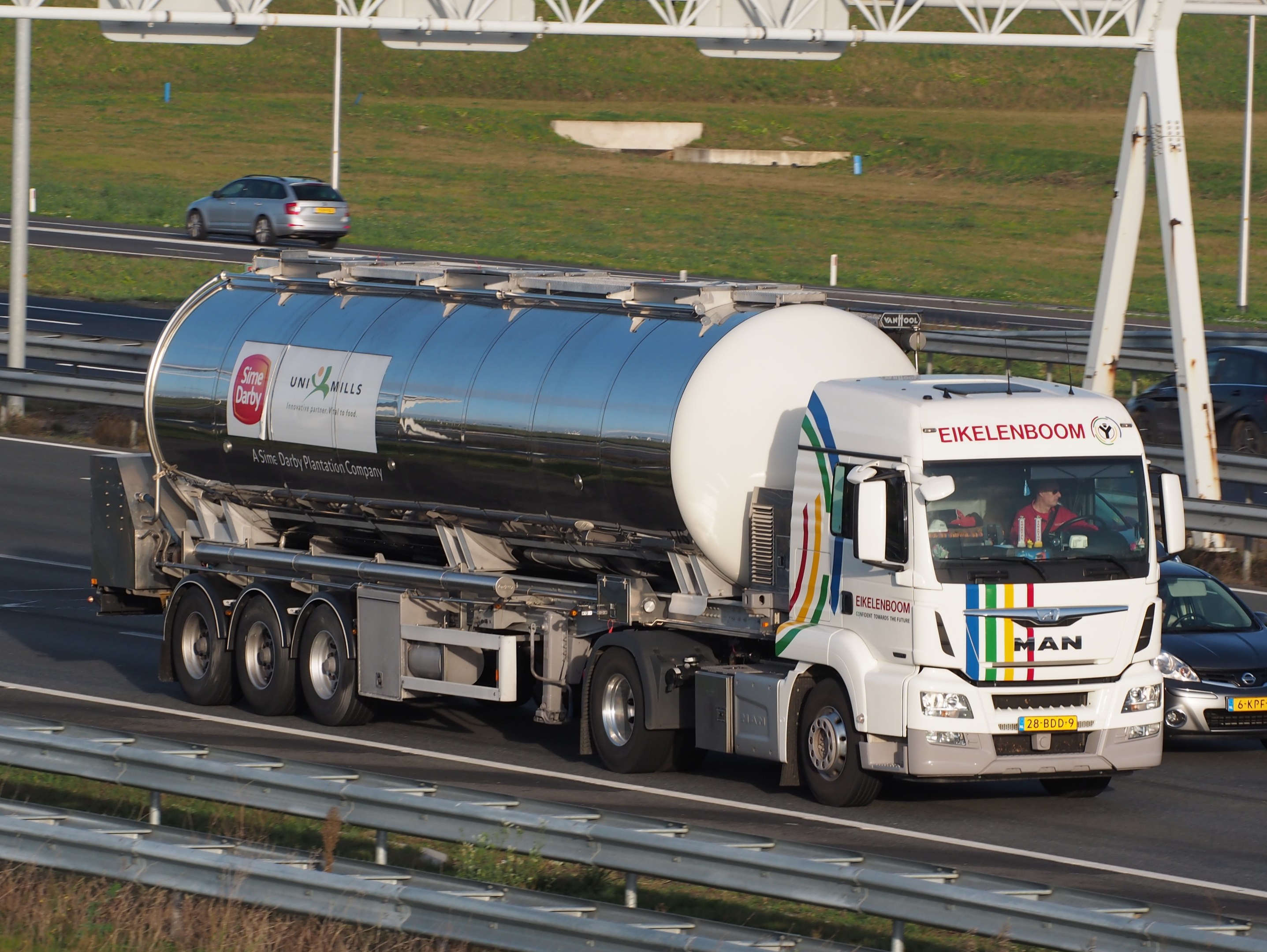 Großer Tanklastwagen auf einer Autobahn neben einem Auto mit sichtbaren Passagieren im Truck und Gras, Strommasten und anderen Gegenständen im Hintergrund.