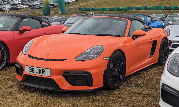 Ein Porsche 718 Boxster GTS auf der Goodwood Festival of Speed, zusammen mit anderen Autos, einem Banner, Bäumen und einem klaren blauen Himmel im Hintergrund.
