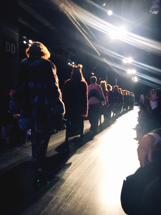 Eine Gruppe von Menschen läuft über den Laufsteg einer Fashion Show, während Zuschauer auf beiden Seiten sitzen.