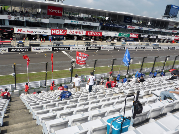 Eine Gruppe von Menschen sitzt auf der Tribüne eines Rennstadions, umgeben von Fahnen, Schildern, Geländern und Stufen, unter einem bewölkten Himmel.