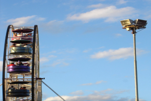 Eine Gruppe von Menschen im Riesenrad mit einem beleuchteten Mast rechts und einem bewölkten Himmel im Hintergrund.
