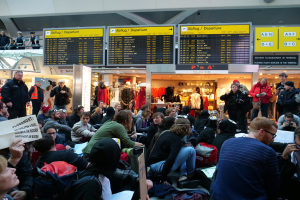 Eine große Gruppe von Menschen, die auf einem Flughafen während einer Protestaktion sitzen und stehen, mit Informationsschildern, Schaufensterpuppen in Kleidern und Deckenbeleuchtung im Hintergrund.