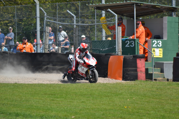 Eine Person in Helm und Handschuhen fährt auf einem Motorrad über ein grasbewachsenes Feld, mit Zuschauern, Absperrung, Treppe und einem nummerierten Schuppen im Hintergrund.