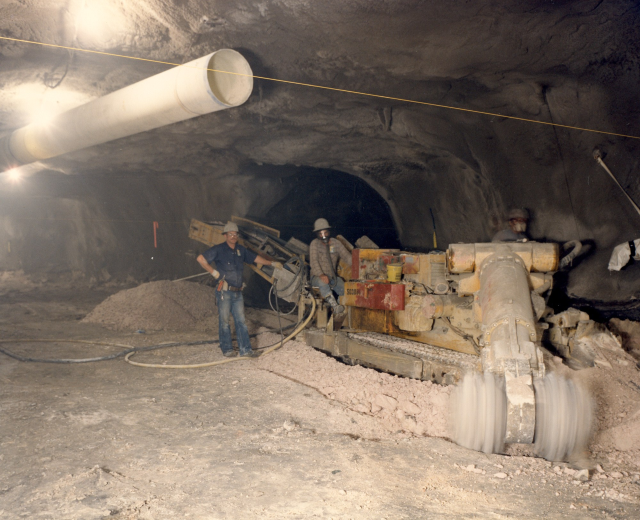Gruppe von M├Ąnnern in Helmen, die in einem dunklen Stollensystem mit Maschinen und Rohren arbeiten.