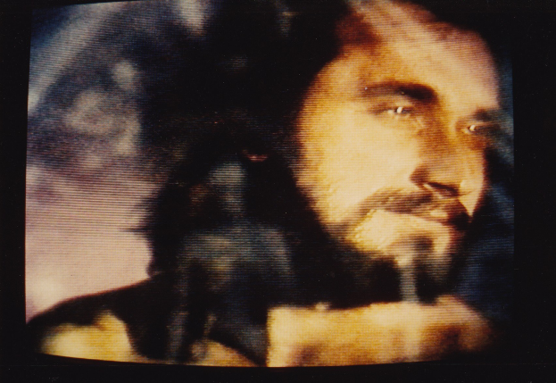 Ein Mann mit langem dunklem Bart und Schnurrbart, der ein weißes Hemd trägt, appears auf einem Fernseher mit dunklem Hintergrund und schaut direkt in die Kamera mit einem ernsten Gesichtsausdruck.