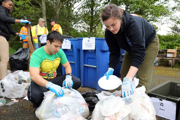 Eine Gruppe von Menschen mit Handschuhen sammelt Müll in einem Park, mit Plastikabdeckungen, Flaschen und anderem Müll auf dem Boden in der Nähe eines Mülleimers und einer hölzernen Bank, vor einem Baum und einem klaren blauen Himmel.