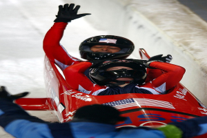 Zwei Bobsleighs im Einsatz bei den Olympischen Spielen, einer mit sichtbarem Helm und Handschuhen, mit einer Person unten und einer Wand im Hintergrund.