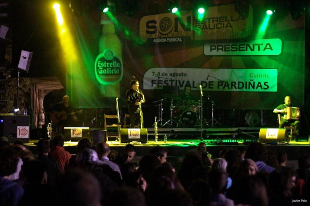 Zuschauende auf einer Bühne mit Musikinstrumenten, Lautsprechern, Mikrofonen, einer Kulisse und Scheinwerfern, mit einer Flasche und anderen Gegenständen im Hintergrund.