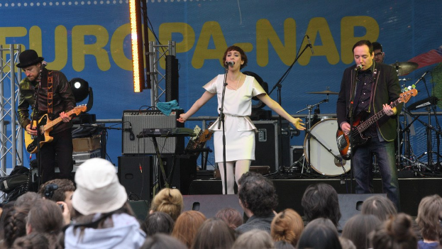 Vier Personen auf der Bühne: eine Frau in einem weißen Kleid, die singt, flankiert von zwei Männern mit Gitarren, mit einer Menge unten und Lautsprechern an der Wand im Hintergrund.