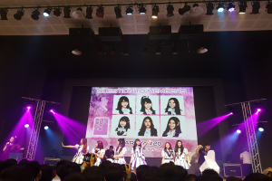 Eine Gruppe von Menschen tritt auf einer hell beleuchteten Bühne mit Trägern auf, während eine Menge darunter das Ereignis aufzeichnet; ein großer Bildschirm im Hintergrund zeigt Bilder der K-Pop-Gruppe BK48.