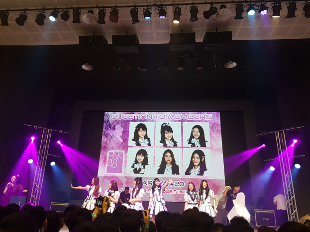 Eine Gruppe von Menschen tritt auf einer hell beleuchteten Bühne mit Trägern auf, während eine Menge darunter das Ereignis aufzeichnet; ein großer Bildschirm im Hintergrund zeigt Bilder der K-Pop-Gruppe BK48.