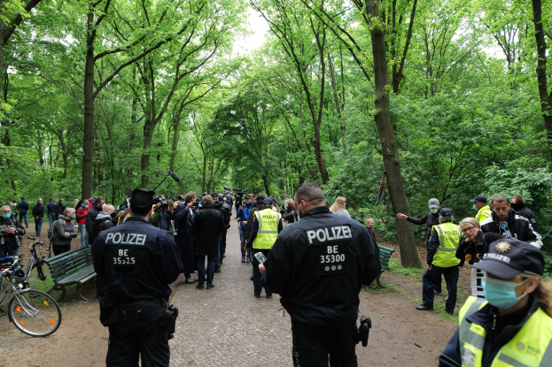 Eine Gruppe Polizisten vor einer Menge während einer Anti-Terror-Demonstration in Berlin, mit Fahrrädern und einer Bank im Vordergrund und Bäumen im Hintergrund.