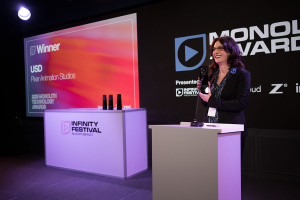 Frau in einem schwarzen Kleid, die an einem Rednerpult steht und einen Preis hält, mit einem Mikrofon und anderen Gegenständen auf dem Pult, vor einem Bildschirm und einer Werbetafel.