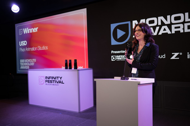 Frau in einem schwarzen Kleid, die an einem Rednerpult steht und einen Preis hält, mit einem Mikrofon und anderen Gegenständen auf dem Pult, vor einem Bildschirm und einer Werbetafel.