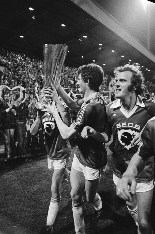 Schwarzes Foto von Fußballspielern, die mit einem Pokal auf einem Feld feiern, umgeben von Zuschauern hinter einem Zaun unter Stadionbeleuchtung.