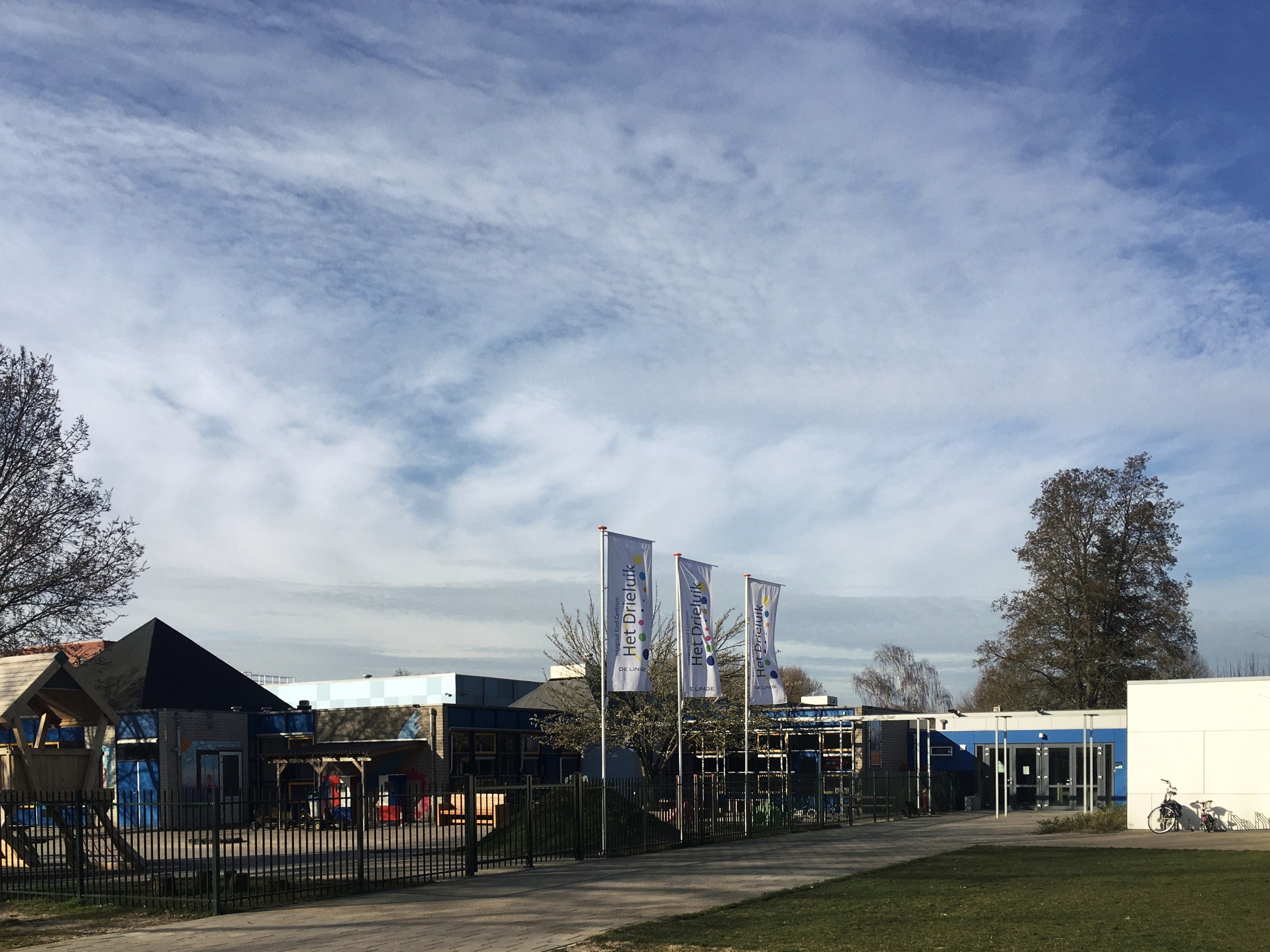 Großes Backsteingebäude mit Metallzaun, Rasenfläche, Weg, Bäumen, Bannern, einem geparkten Fahrrad, nahen Häusern und einem bewölkten Himmel, identifiziert als neuer Spielbereich für Kinder an der Schule.