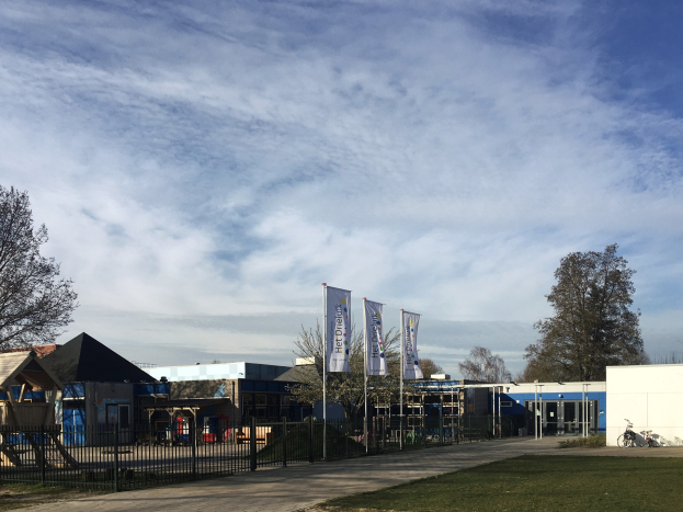Großes Backsteingebäude mit Metallzaun, Rasenfläche, Weg, Bäumen, Bannern, einem geparkten Fahrrad, nahen Häusern und einem bewölkten Himmel, identifiziert als neuer Spielbereich für Kinder an der Schule.