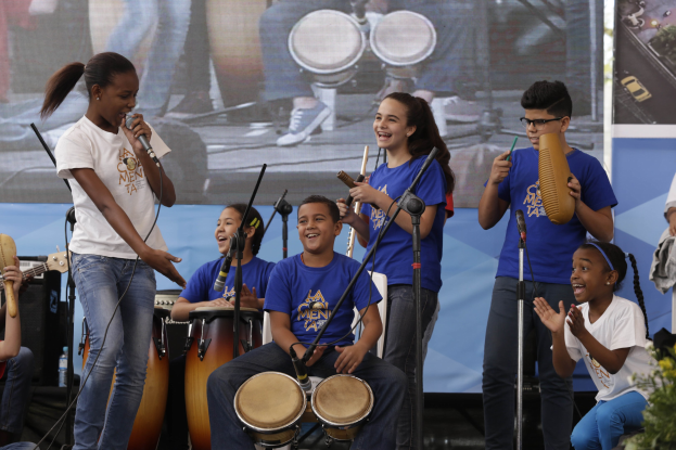 Eine Gruppe von Kindern, die auf einer Bühne Musikinstrumente spielen, mit einer Frau, die ein Mikrofon hält, umgeben von Mikrofonstativen und einem Banner im Hintergrund, mit Pflanzen auf der rechten Seite.