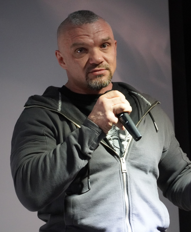 Ein Mann in einer schwarzen Kapuzenjacke und einem schwarzen Anzug mit einem weißen Hemd, der ein Mikrofon hält, steht selbstbewusst vor einer Wand.