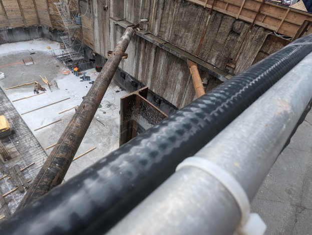 Große Metallrohr an einer Baustelle umgeben von Arbeitern, Holzwerkstoffen und Ausrütung, mit einer Person in einem blauen Hemd rechts und Fahrzeugen und einem Gebäude im Hintergrund.
