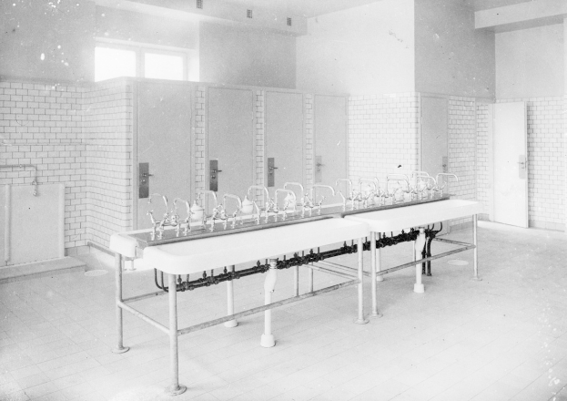 Schwarz-weiß-Foto einer öffentlichen Toilette mit einer Reihe von Waschbecken, Hähnen, Wänden, Türen, Fenstern und einem gefliesten Boden, mit verschiedenen Gegenständen, die herumliegen.