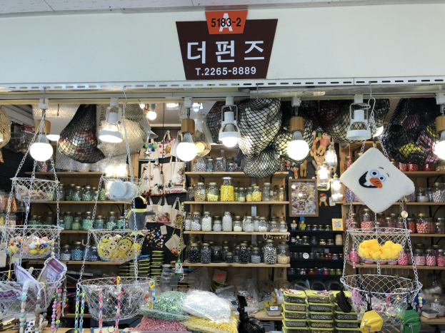 Ein Ladeneinrichtungsbild mit verschiedenen Gegenständen wie Körbchen, Lichtern, Flaschen und Spielzeugen, die auf Regalen angeordnet sind, mit einer Tafel öber dem Eingang, die es als bestes koreanisches Geschäft in Seoul ausweist.
