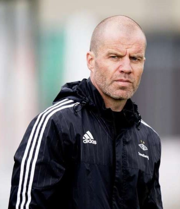 Baldiger Trainer in marineblauer Jacke mit weißen Streifen und weißem Kragen, der auf einem Fußballfeld steht und die Hände gefaltet hat.