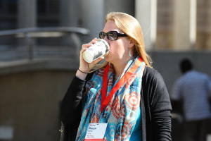 Eine Frau mit Schutzbrille und einem Ausweis um den Hals trinkt aus einem Becher, mit einem unscharfen Hintergrund.