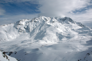 Ein schneebedeckter Berg mit ein paar Skifahrern, die hinunterfahren, umgeben von einem bewölkten Himmel und unberührtem Schnee.