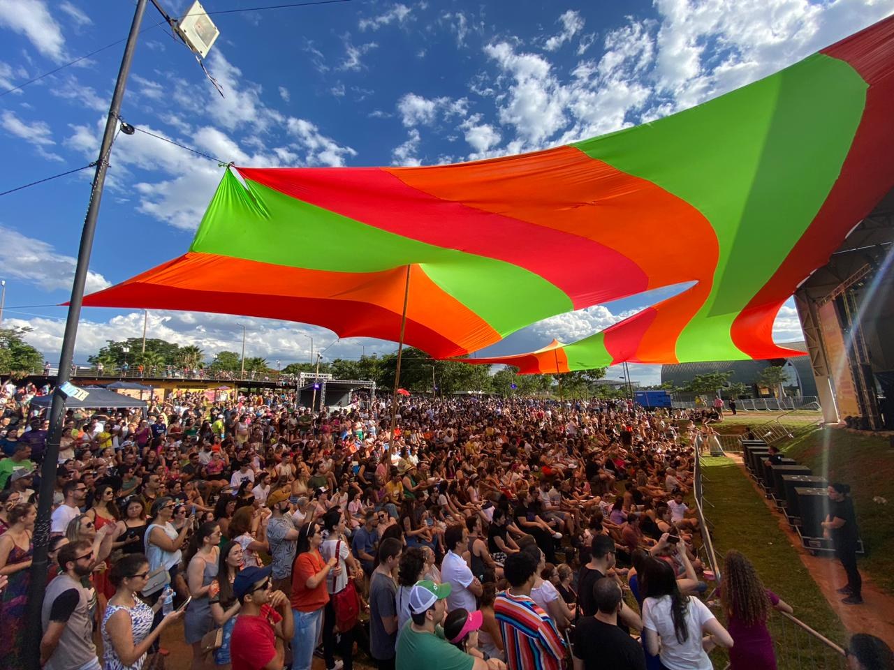 Eine gro%C3%9Fe Menge steht vor einer Regenbogenflagge mit einem Geb%C3%A4ude, Lautsprechern auf der B%C3%BChne, B%C3%A4umen und Wolken im Hintergrund bei einer Festival-Jubil%C3%A4umsfeier.