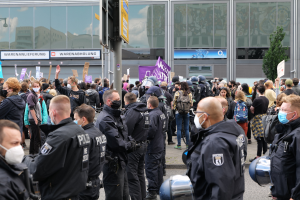 Große Menschenmenge protestiert vor einem Gebäude, einige halten Schilder und tragen Helme, mit einem Schildständer und einem Baum im Vorder- und Hintergrund.