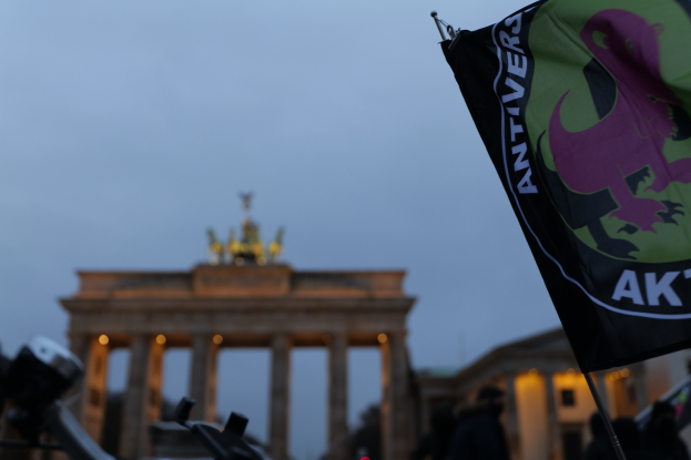 Eine Gruppe von Menschen steht vor dem Brandenburger Tor in Berlin, Deutschland, mit einer Fahnenstange mit Logo und Text im Vordergrund.