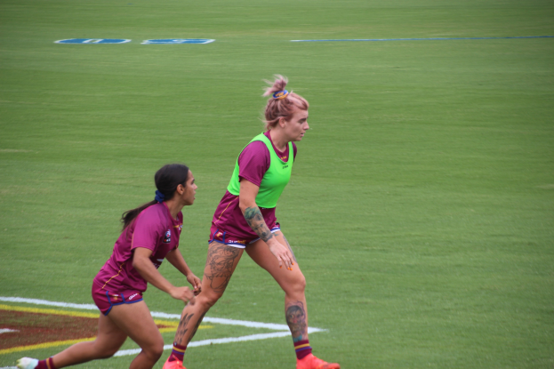 Zwei Frauen in Sportbekleidung spielen Fußball auf einem Feld, eine in einem grünen T-Shirt und Shorts, die andere in einem pinken Kleid, beide mit sichtbaren Tattoos an Händen und Füßen.