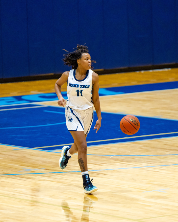 Eine Frau in einer blauen und weißen Uniform dribbelt einen Basketball auf einem Court, trägt ein weißes T-Shirt mit der Aufschrift "Wake Tech Women's Basketball" und blaue Schuhe, mit einer blauen Wand im Hintergrund.