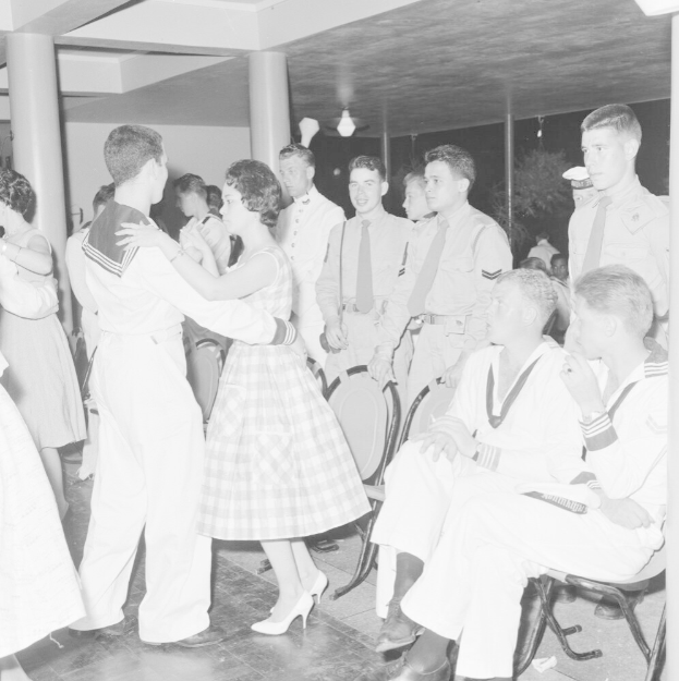 Schwarz-weißes Bild einer Gruppe von Menschen in Militäruniformen, die auf einem Tanzboden tanzen und sitzen, mit Säulen und Lichtern im Hintergrund.