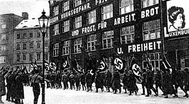 Ein Schwarz-Weiß-Bild einer Gruppe von Menschen, die eine Straße entlangmarschieren, mit Fahnen und Transparenten in den Händen, mit einem Laternenmast links und einem Gebäude mit einer Tafel mit der Aufschrift "Deutsche Nazi-Partei in Berlin" rechts.