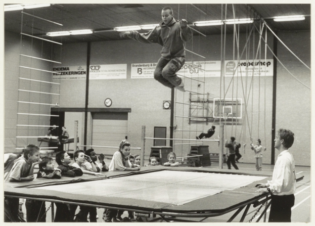 Kinder sitzen um ein Trampolin in einer Turnhalle mit einer Person in der Luft, einem Jungen daneben, einem Tisch unten und Hintergrund-Elementen wie Menschen, einer Uhr, Texttafeln und Deckenleuchten.