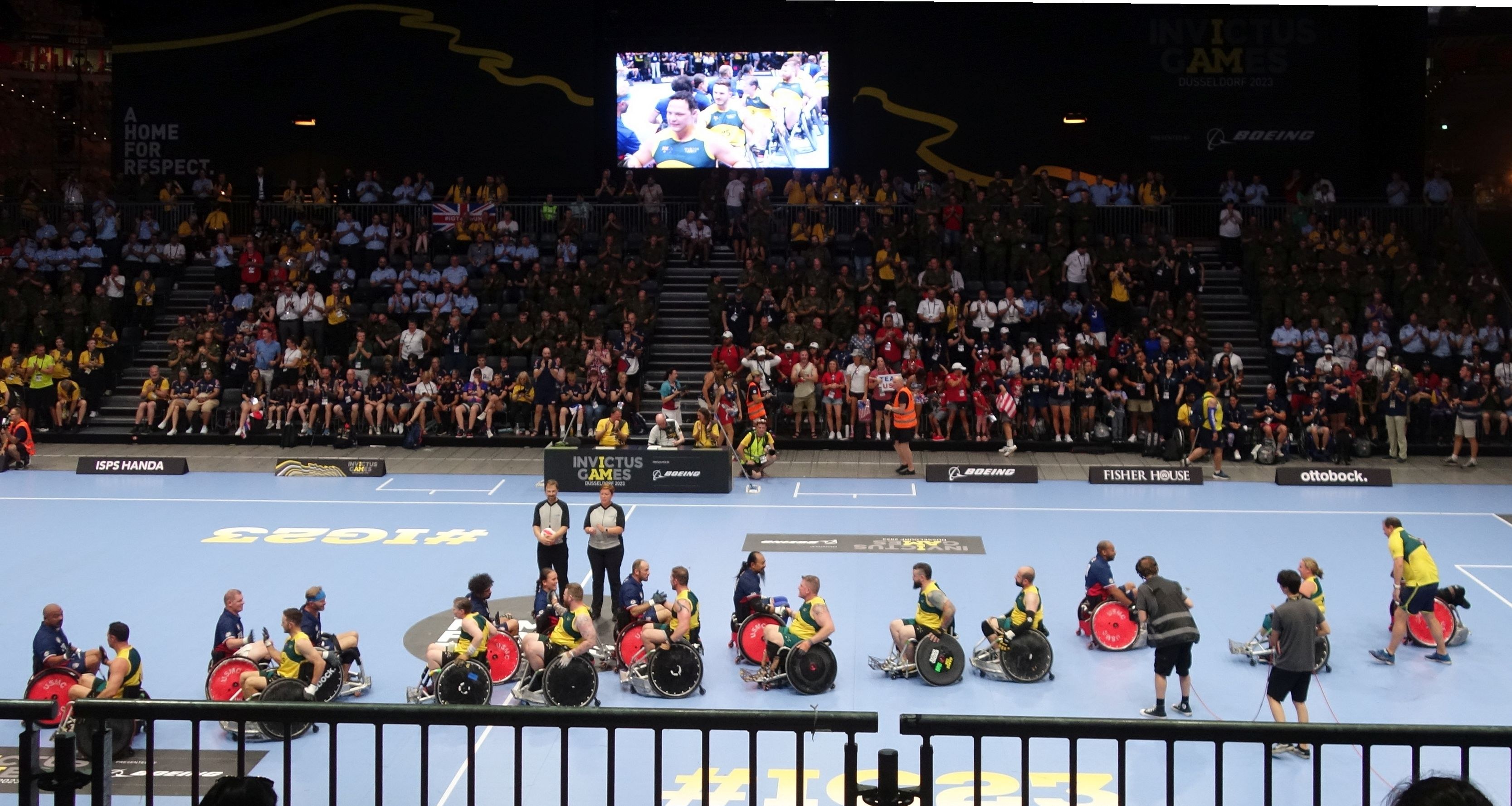 Eine Gruppe von Menschen im Rollstuhl auf einem Platz bei den Invictus Games Düsseldorf 2020, umgeben von einer Absperrung und einer großen Zuschauermenge, mit Stufen zu einem Bildschirm und Informationsschildern im Hintergrund.