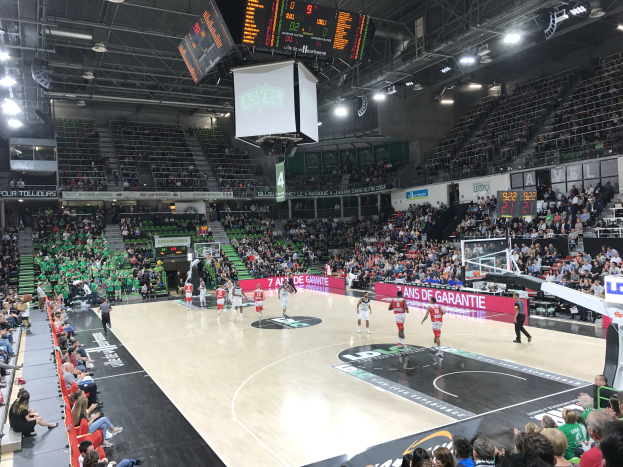 Basketballspiel in einer großen Arena mit Zuschauern, mit "Asvel vs Galatasaray"-Anzeigetafeln auf dem Platz.