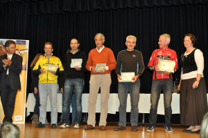 Eine Gruppe von Menschen auf einer Bühne, die Zertifikate halten, mit einem Banner im Hintergrund, einem Tisch zur Seite und einem schwarzen Vorhang.