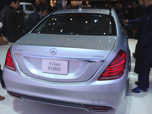 Silberner Mercedes S-Klasse Hybrid auf einer Automesse präsentiert, umgeben von Menschen in verschiedenenfarbigen Kleidern, mit anderen Fahrzeugen, Tafeln und Beleuchtung im Hintergrund.