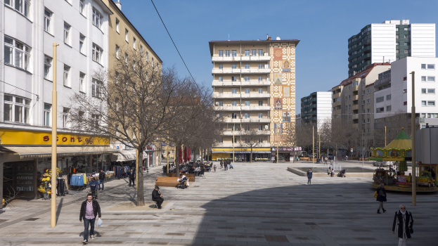 Ein belebter Stadtplatz mit Menschen, die spazieren gehen, auf Bänken sitzen und stehen, umgeben von Bäumen, Gebäuden und einer Karussell im Hintergrund unter einem sichtbaren Himmel.