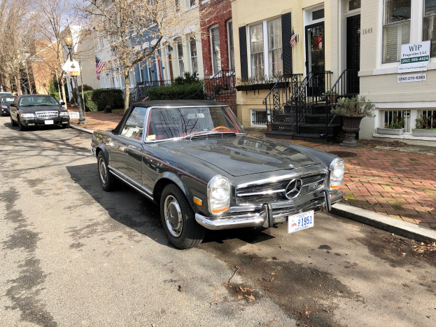 Ein 1971 Mercedes-Benz 280SL, der auf einer Stra├če neben Geb├Ąuden mit Fenstern, Treppen, Topfpflanzen, B├Ąumen, Laternenmasten und Texttafeln unter einem sichtbaren Himmel geparkt ist.