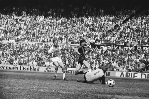 Schwarzes und weißes Foto von Fußballspielern im Einsatz auf einem Feld mit Zuschauern im Hintergrund und einer Bundesliga 1978-79 Ajax vs. Hannover 96-Tafel.